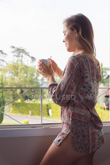 Продумана блондинка в сорочці з чашкою кави, яка дивиться у вікна вранці — стокове фото