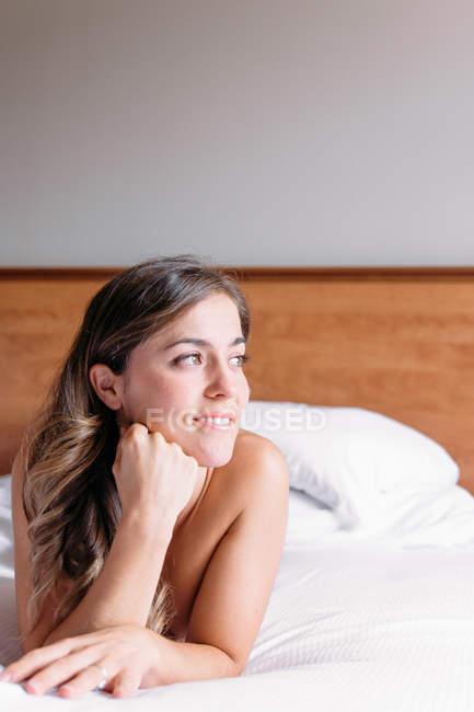 Una chica rubia sonriente en una cama con sábanas blancas iluminadas por la luz de la ventana por la mañana. - foto de stock