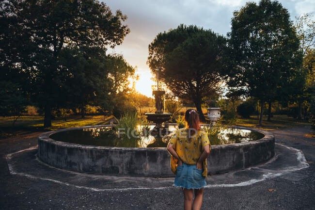 Back view of bronzed woman relaxing near aged fountain and looking at sunset in summer park — Stock Photo