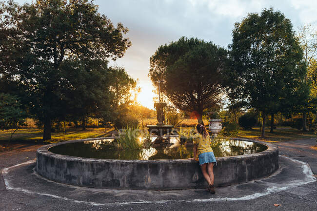 Vue arrière de la femme bronzée se détendre appuyé sur la fontaine et regardant le coucher du soleil dans le parc d'été — Photo de stock