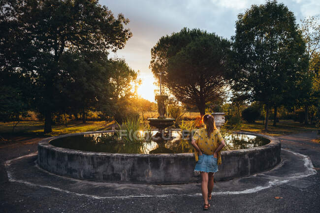 Visão traseira da mulher bronzeada relaxante perto da fonte envelhecida e olhando para o pôr do sol no parque de verão — Fotografia de Stock