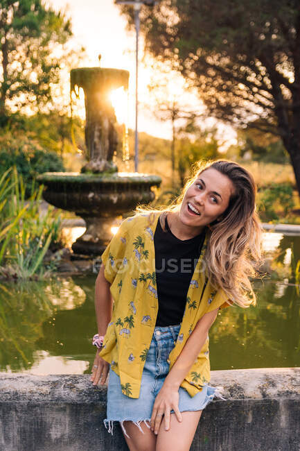 Affascinante donna sorridente in abbigliamento casual appoggiata alla fontana al tramonto e guardando la fotocamera retroilluminata — Foto stock