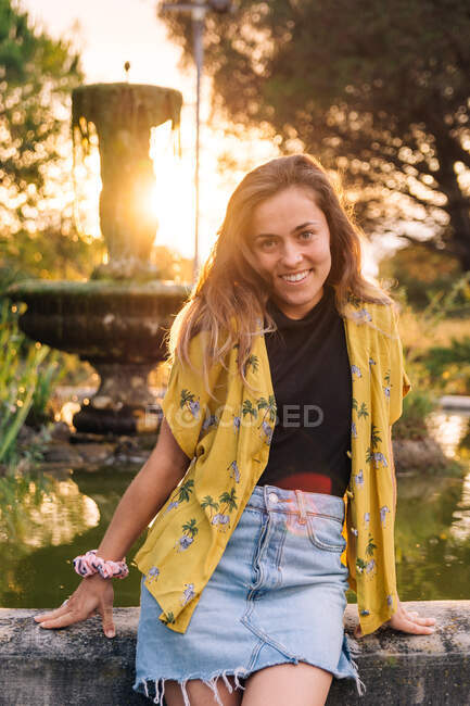 Mulher sorridente encantadora em desgaste casual inclinado na fonte ao pôr do sol e olhando para a câmera na parte traseira iluminada — Fotografia de Stock