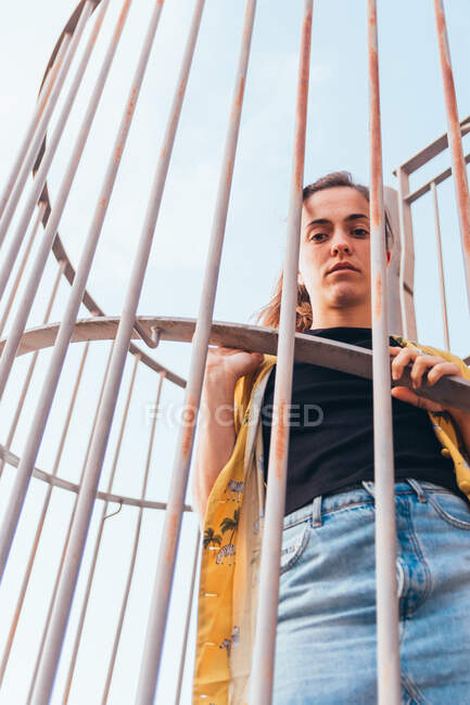 From below sensual casual woman standing in cage on clear sky in summer and looking at camera — Stock Photo