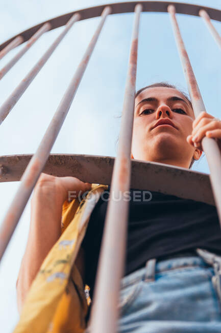 Vista de cerca desde abajo sensual mujer casual de pie en la jaula en el cielo despejado en verano y mirando a la cámara - foto de stock