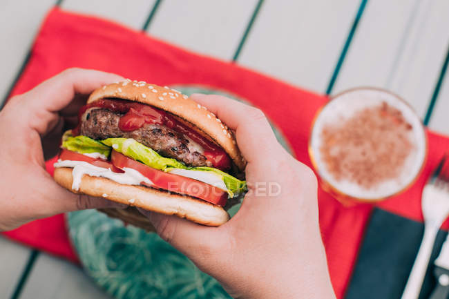 Manos de persona sosteniendo deliciosa hamburguesa casera de queso con lechuga, tomate y salsa . - foto de stock