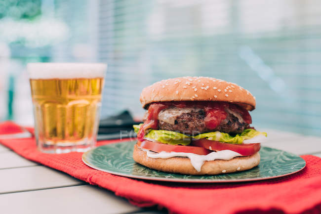 Вкусный домашний гамбургер с салатом, помидорами и соусом на зеленой тарелке, подаваемый со стаканом пива . — стоковое фото