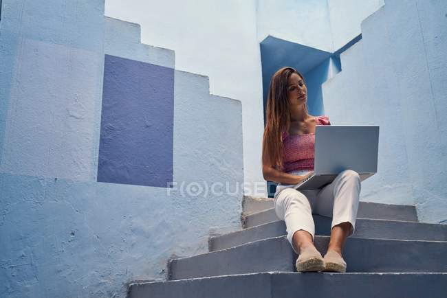 Прекрасна жінка сидить на блакитних сходах синього будинку і користується комп 