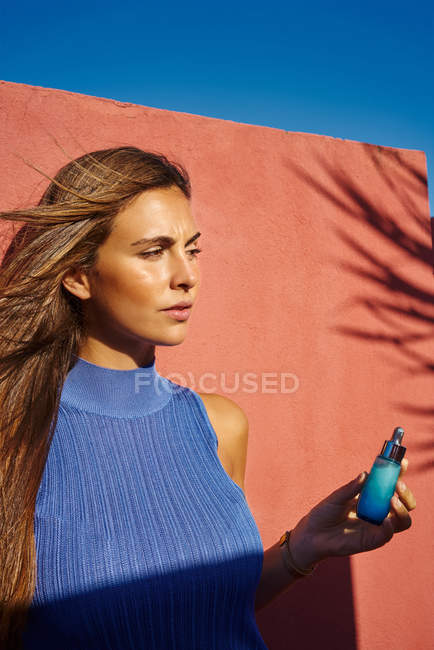 Красива жінка в блакитній сукні, спираючись на будівлю червоних стін, тримає косметичну пляшку — стокове фото