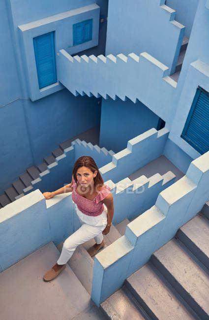 Вид зверху на молоду жінку, що йде нагору на сучасній блакитній будівлі і дивиться на камеру — стокове фото