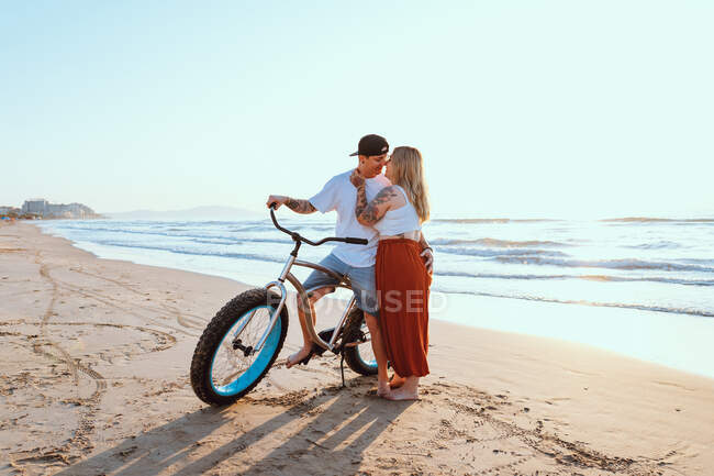 Tender casal moderno abraçando e de pé com bicicleta à beira-mar de verão — Fotografia de Stock