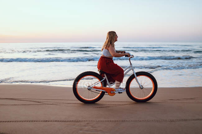 Vista lateral de la gordita mujer feliz moderna en la falda brillante montar en bicicleta a lo largo de la costa del océano escénico en crepúsculo - foto de stock