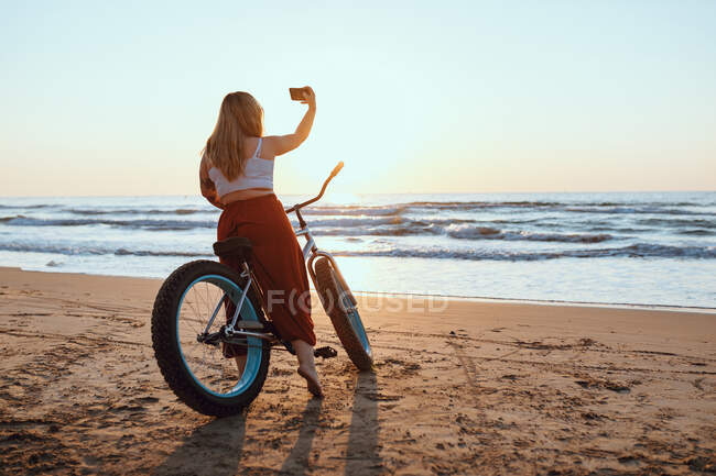 Cheerful chubby woman taking selfie on smartphone while resting with bicycle at sunny shore — Stock Photo