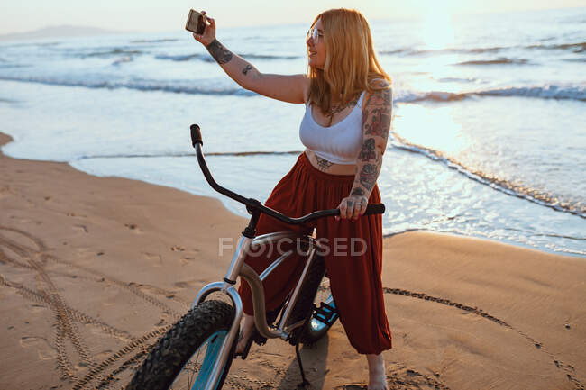 Mulher gordinha alegre tomando selfie no smartphone enquanto descansa com bicicleta na costa ensolarada — Fotografia de Stock