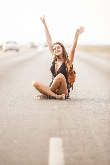 Jolie jeune femme souriante et assise sur la route avec des rayures blanches et regardant la caméra avec les mains levées — Photo de stock