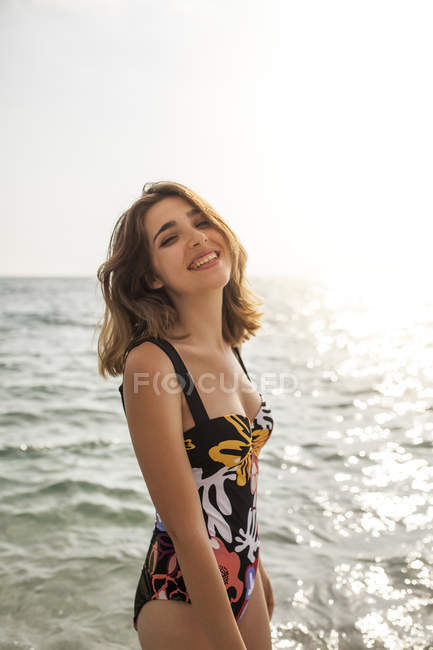 Привлекательная счастливая женщина в стильном купальнике стоит и смотрит в камеру в солнечный день — стоковое фото