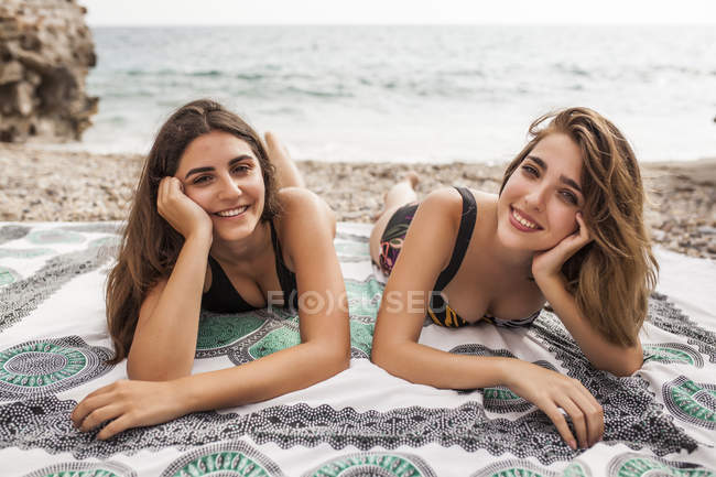 Zwei junge Frauen in Badebekleidung, die es sich auf einer Decke am Strand bequem machen und in die Kamera schauen — Stockfoto