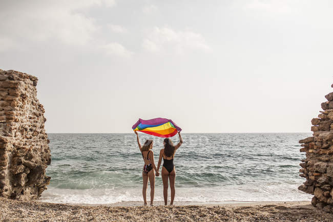 Вид на лесбийскую пару, стоящую на пляже с красочным флагом движения ЛГБТ во время летних каникул — стоковое фото