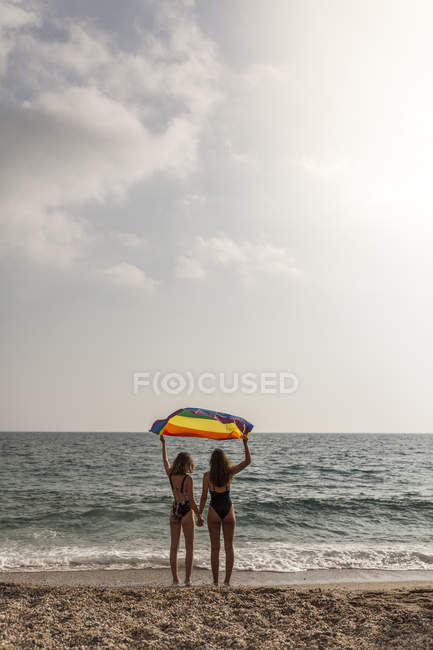 Вид на лесбийскую пару, стоящую на пляже с красочным флагом движения ЛГБТ во время летних каникул — стоковое фото