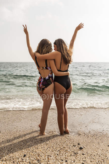Back view of young women in swimsuits standing by sea, embracing with raising hands — Stock Photo