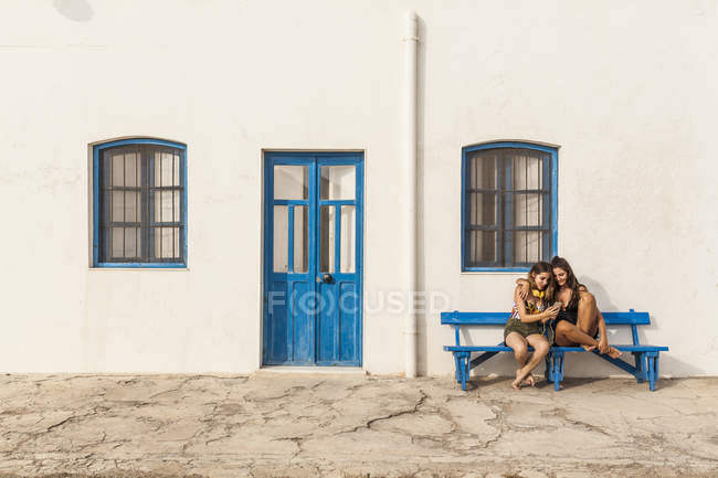 Happy young women in tops and shorts sitting on bench and using cellphone — Stock Photo