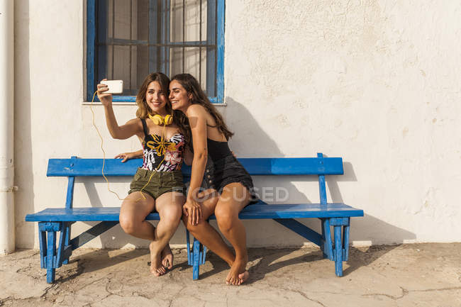 Mujeres jóvenes felices en tops y pantalones cortos sentados en el banco, abrazando y tomando selfie en el teléfono inteligente - foto de stock