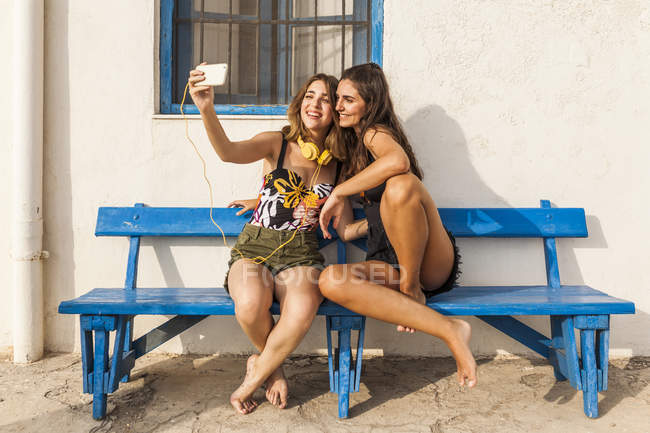 Happy young women in tops and shorts sitting on bench, embracing and taking selfie on smartphone — Stock Photo