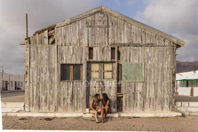 Affettuosa coppia femminile seduta vicino al vecchio capanno di legno, che si tiene per mano e si tocca con la fronte — Foto stock