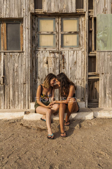 Affettuosa coppia femminile seduta vicino al vecchio capanno di legno, che si tiene per mano e si tocca con la fronte — Foto stock