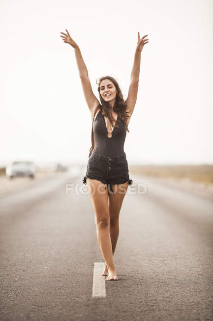 Привлекательная молодая босиком женщина улыбается и ходит по пустой дороге с рюкзаком, поднимая руки и глядя в камеру — стоковое фото