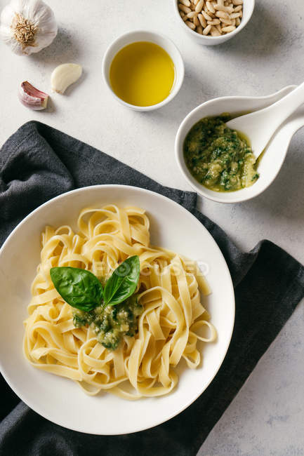 Top view of white plate with fresh cooked tagliatelle pasta with pesto sauce next to garlic and oil on kitchen table — Stock Photo