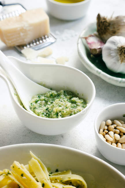 Colorful fresh pesto sauce in bowl next to garlic and cheese with pine nuts on rustic white table — Stock Photo