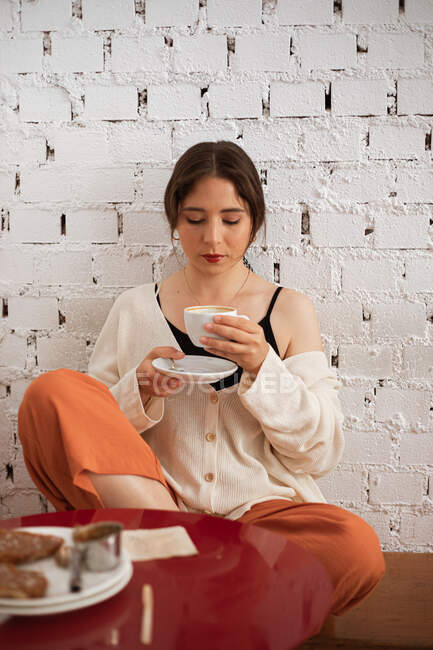 Расслабленная женщина в домашней одежде пьет из кружки, сидя за кухонным столом с едой у стены белого кирпича — стоковое фото