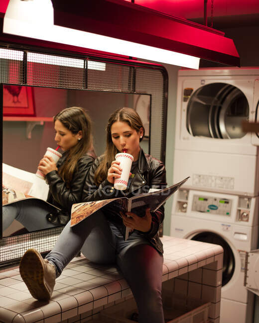 Confident teen woman in leather jacket drinking with straw from takeaway cup and reading magazine while waiting for laundry done in public laundromat — Stock Photo