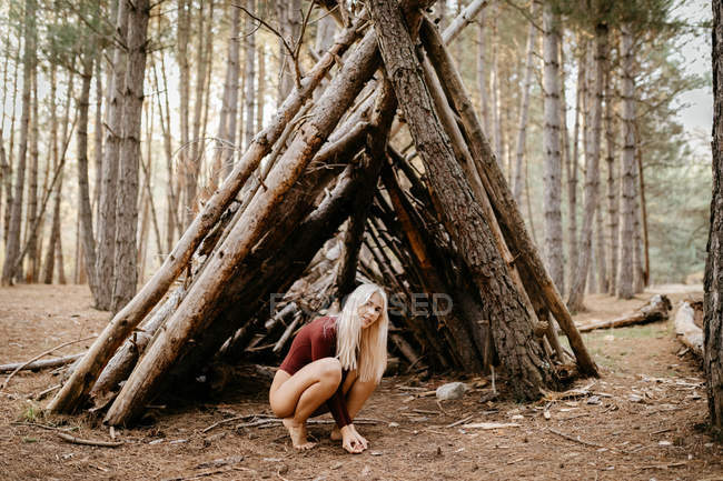 Vista laterale della donna spensierata bionda a piedi nudi in body luminoso seduta su hunkers accanto alla capanna nella pineta — Foto stock
