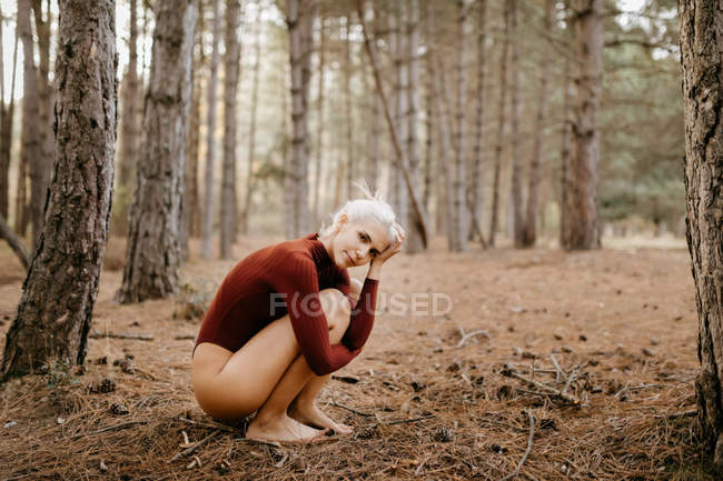 Side view of attractive blonde woman in leotard squatting and embracing knees sitting barefoot in pine forest — Stock Photo