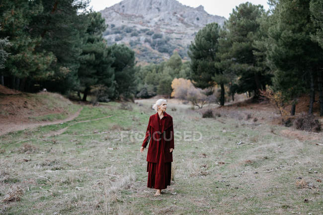 Босоногая блондинка в красном пальто прогуливается по соснам в холодный день — стоковое фото