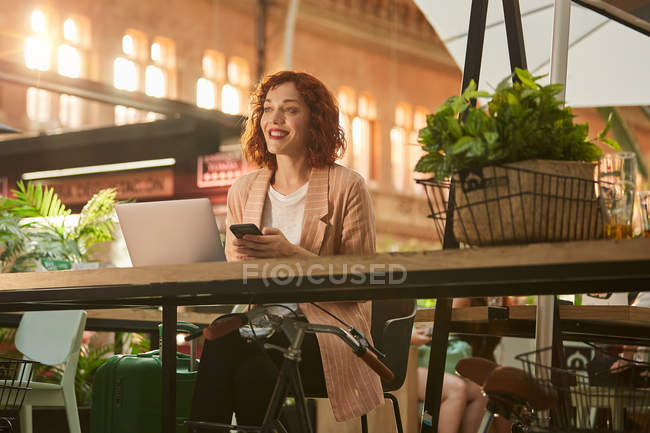Веселая молодая женщина работает на ноутбуке в кафе и просматривает телефон — стоковое фото