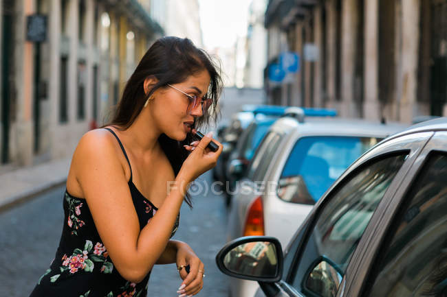 Seitenansicht einer unbeschwerten Frau in Sonnenbrille und Kleid, die beim Anblick des Autofensters hellen Lippenstift aufsetzt — Stockfoto