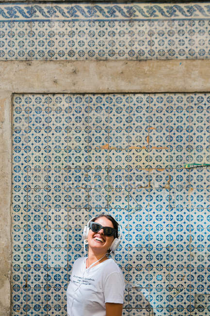 Carefree beautiful woman in casual outfit and headphones standing on blue mosaic wall of building on city street — Stock Photo