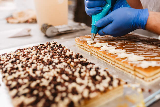 Cozinheiro anônimo espremendo massa de pastelaria fresca na bandeja com papel enquanto trabalhava no fundo borrado da padaria — Fotografia de Stock