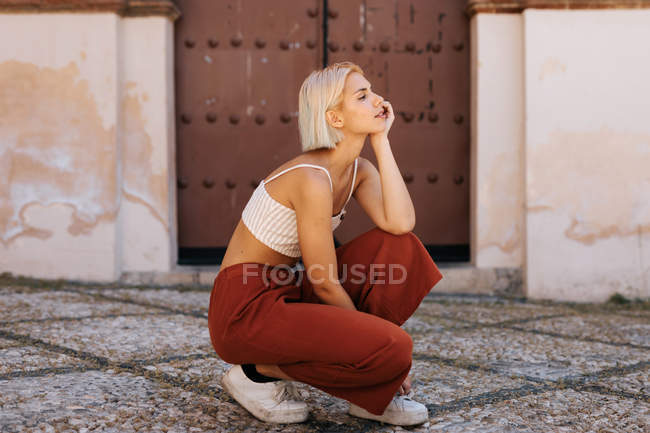 Attrayant jeune femme en tenue élégante accroupi et regardant loin contre l'ancien bâtiment avec portail minable — Photo de stock
