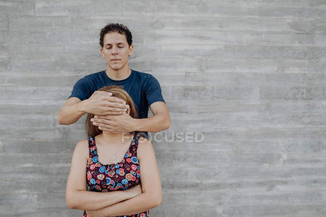 Homem fechar os olhos da mulher em vestido florido de pé nas proximidades parede cinza — Fotografia de Stock
