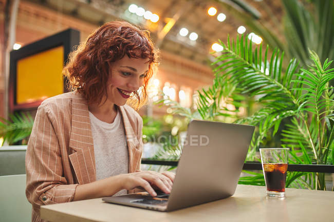Heureuse jeune femme d'affaires assise à table avec une boisson froide dans un café et utilisant un ordinateur portable pour le travail — Photo de stock