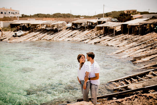Casal alegre em camisas brancas descansando por baía no verão e olhando um para o outro de pé em abraço próximo — Fotografia de Stock