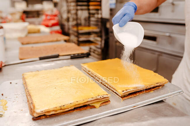 Cocinero anónimo derramando azúcar en polvo de la pala encima de la torta sabrosa mientras trabaja en la cocina de la panadería - foto de stock