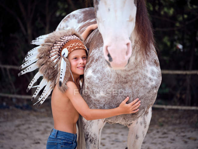 Niño tranquilo mirando hacia otro lado, usando el tradicional sombrero de guerra indio, vinculación con semental de caballo sobre un fondo borroso - foto de stock