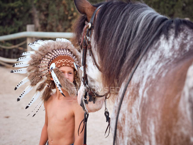 Delighted kid in Indian feather war bonnet with horse on ranch — Stock Photo