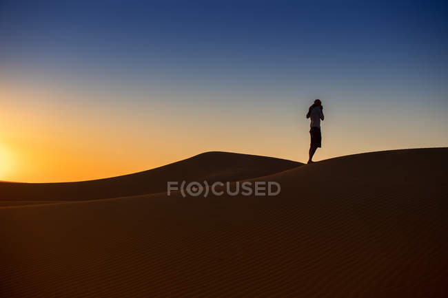 Vista lateral de un turista irreconocible con brazos extendidos que se levantan contra el brillante cielo sin nubes que se hunde en el desierto. - foto de stock