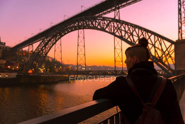 Vista trasera de la turista de pie cerca del terraplén de la ciudad cerca del puente mirando hacia otro lado durante la puesta de sol en Oporto, Portugal - foto de stock